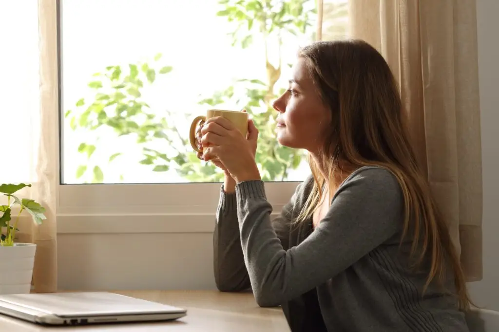 A young woman holding a cup of coffee as she contemplates and looks out of a window.