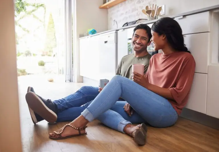 A young couple sitting on their kitchen floor together happily talking to each other.