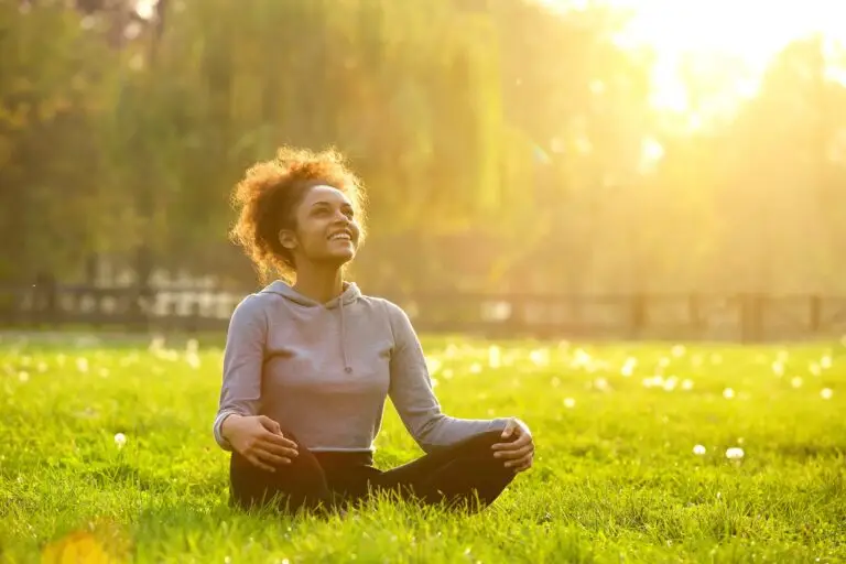 young black woman sitting in the grass at a part in lotus pose, legs crossed, sun shining through the trees as she smiles