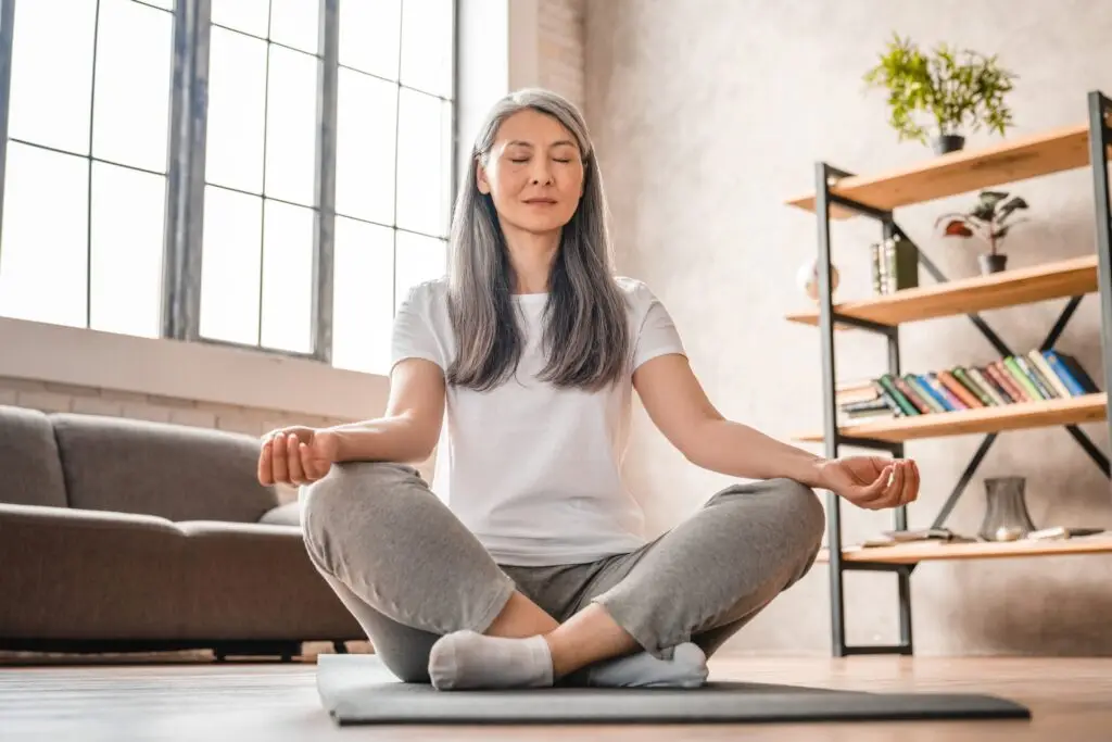 middle aged asian woman with long grey hair sitting on the floor of her living room in lotus pose to meditate.