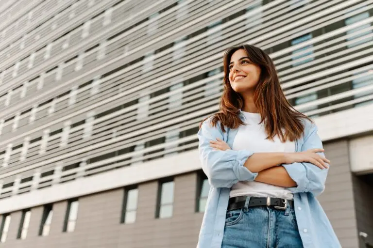 woman standing outside of her workplace in jeans and a long sleeved buttoned shirt, feeling confident and looking off into the distance