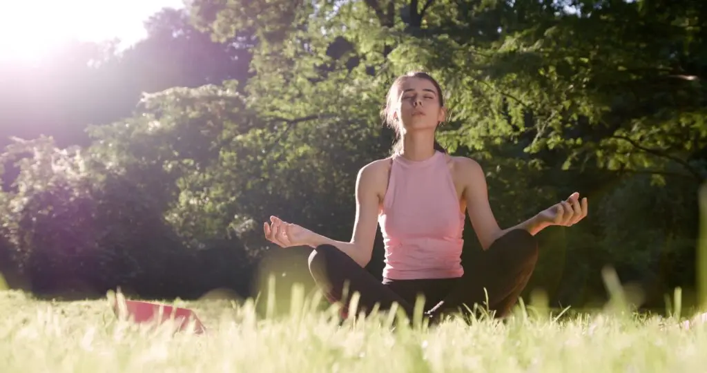 young woman meditation in a field at a park, sitting cross-legged on the ground, elbows on her knees and thumbs connecting to her fingers while she starts to meditate to manage her stress in a healthy way