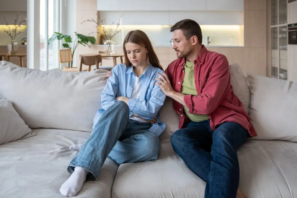 A woman and man sitting on the couch. The man is holding the woman in an aggressive and unwanted way while he gaslights her.