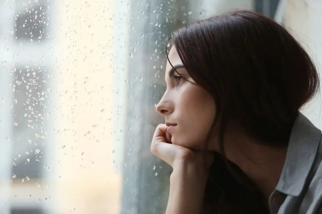 A young brunette woman staring out of a rain-soaked window wondering why she is so unhappy.