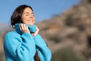 woman standing outside in the fresh air holding the neck portion of her sweater, smiling with her eyes closed as the sun hits her face