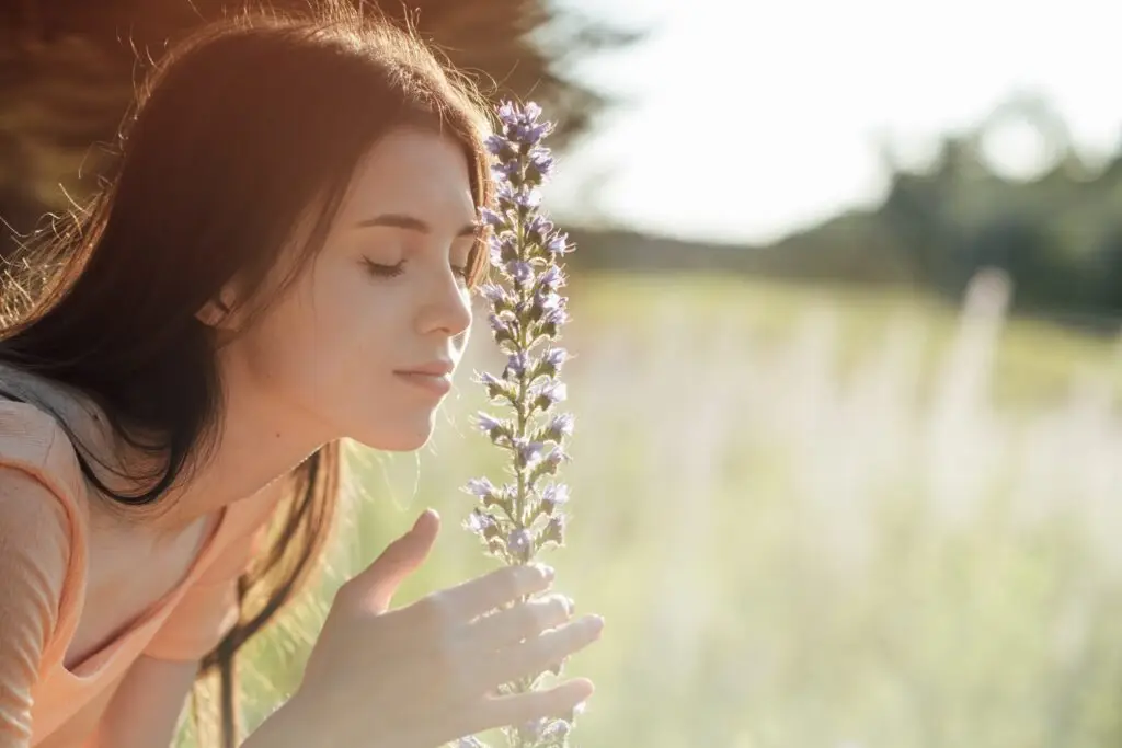 woman outside in a field pulling close a flower with her right hand, closing her eyes and smelling the sweet scent of nature.