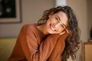 a young woman with dark curly hair smiling wide, leaning to the side, holding her head in her hands.