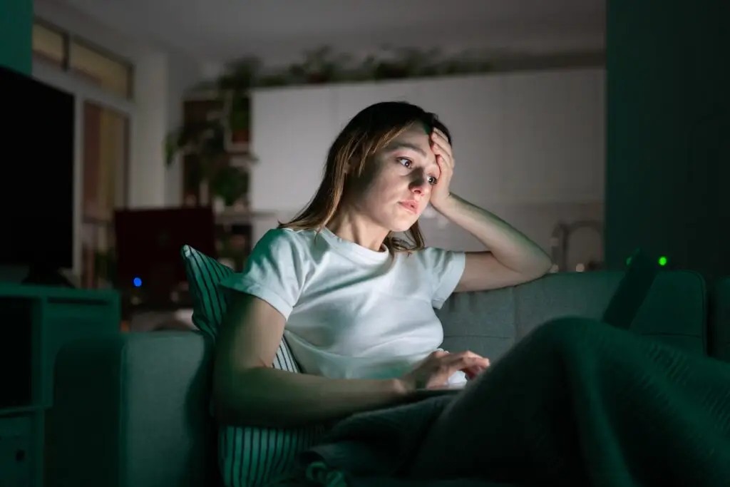 A tired, young, blonde woman sitting on her couch in the dark, on her laptop because her ADHD is causing sleep issues