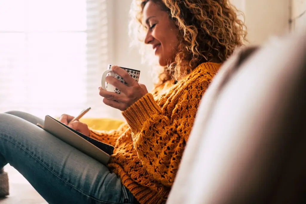 Happy middle-aged woman sitting on her couch sipping tea and writing in her journal about what she wants her life to look like after a divorce.