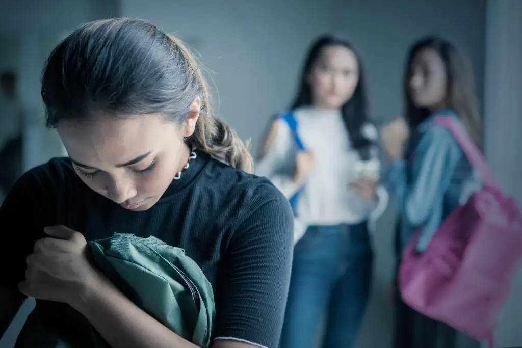 Teenage girl looking down and holding her backpack as her classmates are peer pressuring her in the background.