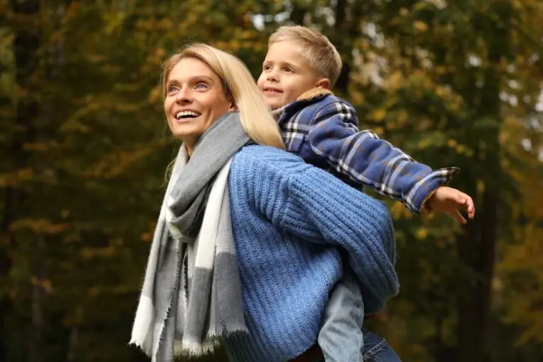 Woman walks around outside with her son on her back. His arms are open wide, enjoying the moment as they spend time outdoors