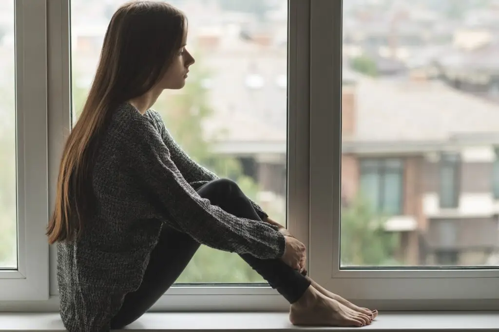 Young woman sitting in her window sill with her knees to her chest, sad with depression over college summer break