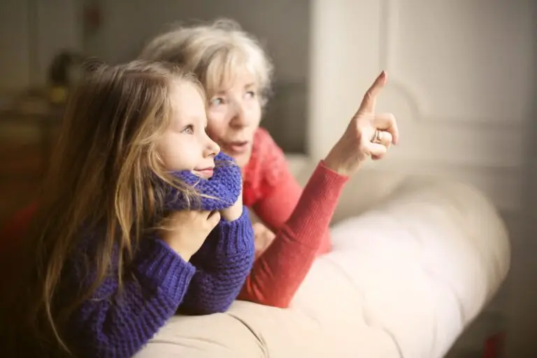 A grandmother spending time with her young granddaughter by crouching together on the back of a couch to look outside of a window together.