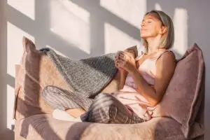 Happy dreamy middle aged woman sitting on sofa in living room with cup of tea or coffee