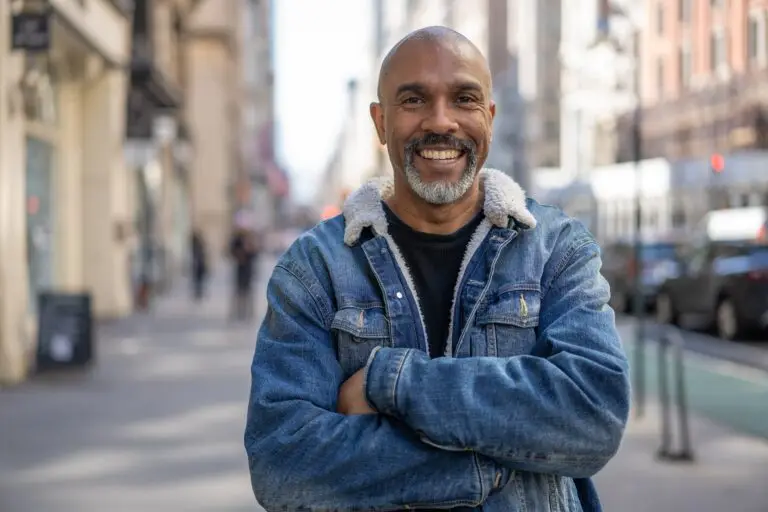 a middle aged African American man standing in public with his arms crossed, smiling wide because he has learned to cope with issues in his life despite societal pressures to keep things to himself.