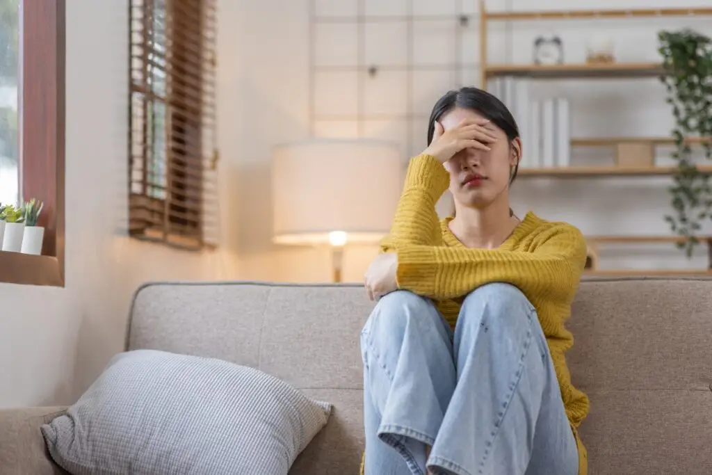 A young woman sitting on her couch with her knees to her chest, elbows resting on her knees, her head resting in one of her hands as she thinks about losing a loved one and all the memories she won't experience with them.