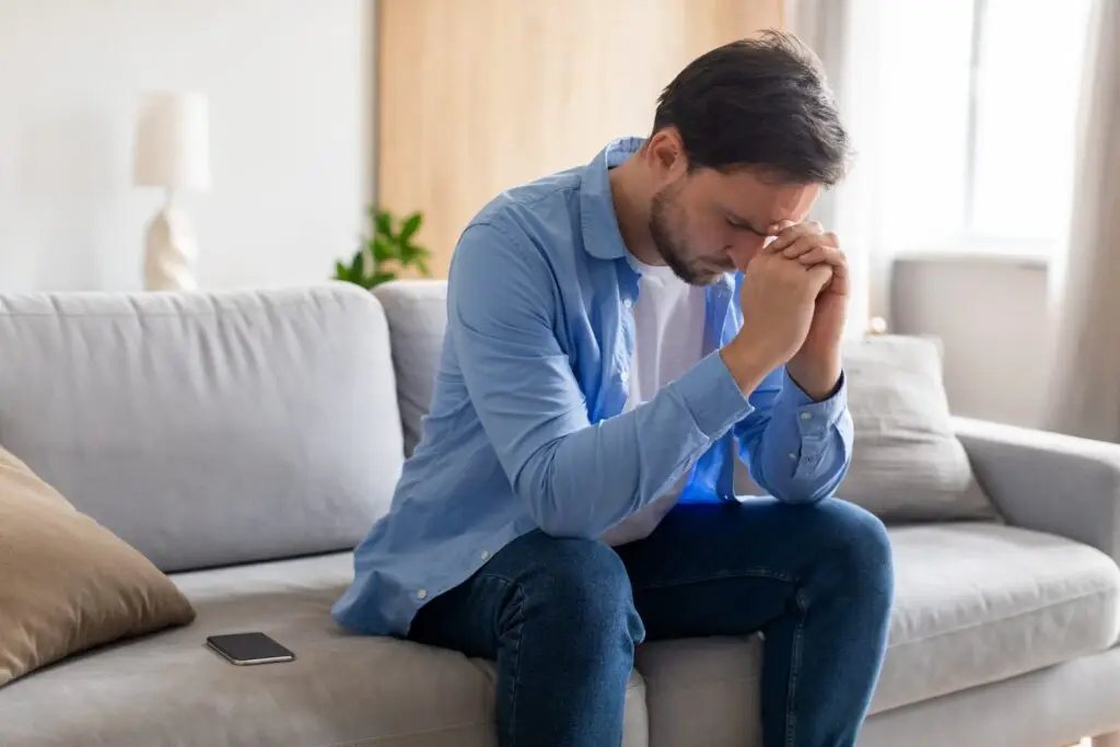 Young man in his late 20's sitting on the couch, elbows rested on his thighs, hands clasped and pressed against his forehead because of troubles he feels he can't talk about with others.