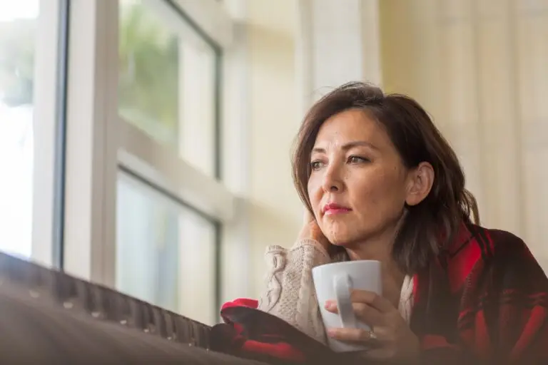 Middle aged woman holding a cup of tea looking out of her window thinking about losing a loved one