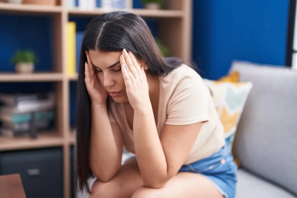 young brunette woman sitting on the edge of her couch, massaging her temples with both hands because she is recognizing how stressed she is while trying to find a new job