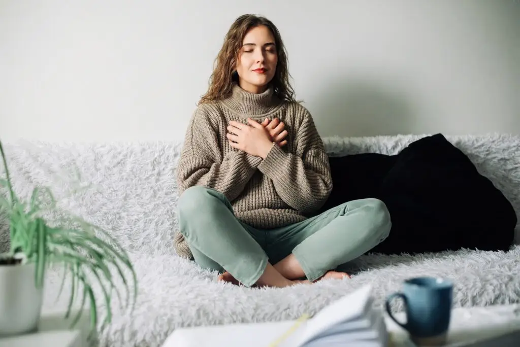 young brunette woman sitting cross-legged on her fuzzy couch, hands crossed over her chest as she takes a deep breath, closes her eyes, and meditates