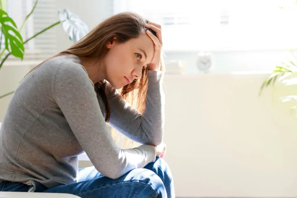 A young woman leaning forward while sitting on a couch. Her hand is holding her forehead while she thinks about how to get a diagnosis for separation anxiety disorder.