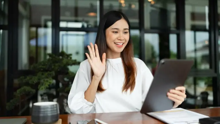 a young brunette woman sitting at her desk at home waving at her online counselor while they video chat on her tablet.