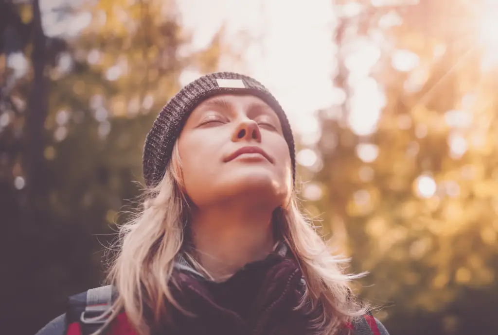 Young blonde woman standing in a wooded area with sunshine and trees, closing her eyes, taking a deep breath, and reframing her thoughts to more positive ones.