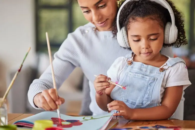 A mother has her young daughter sitting in her lap as they both paint. The young girl has headphones on to reduce sensory input, as she has autism and is sensitive to sounds.