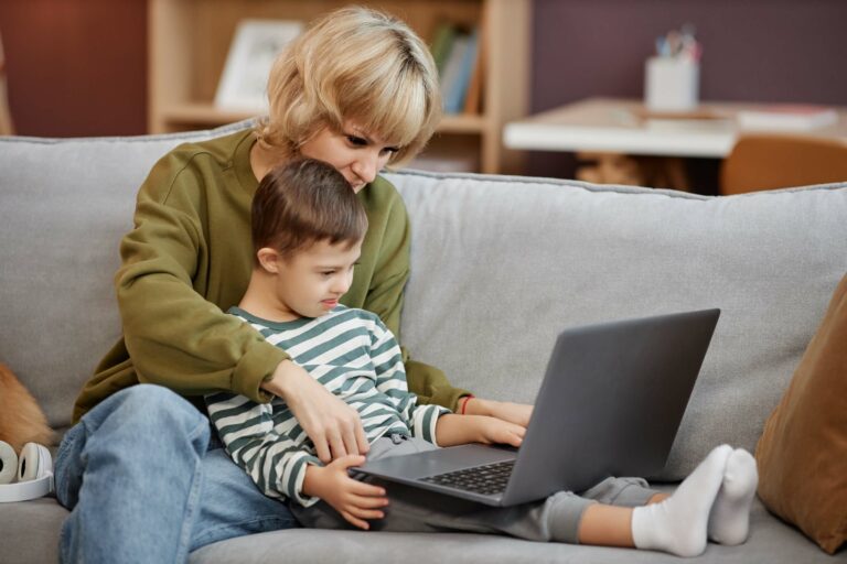 A mother is holding her son in her lap on the couch as they both participate in his online autism therapy with Makin Wellness.