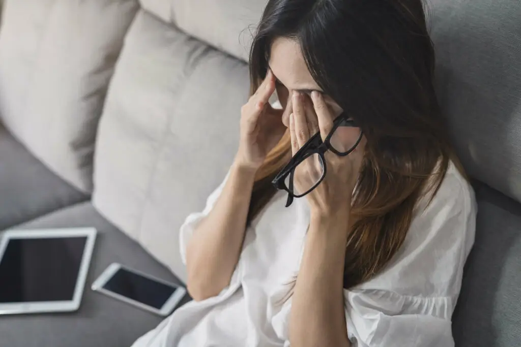 Young woman sitting on her couch, hands rubbing her temples and forehead because of the stress she has over her depression and how it affects her hygiene routine.