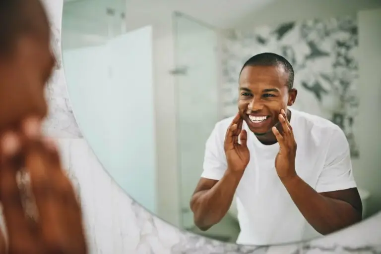 A middle-aged man happy to be taking care of himself with a manageable hygiene routine despite his depression symptoms. He is washing his face while he looks at himself in the mirror, smiling.