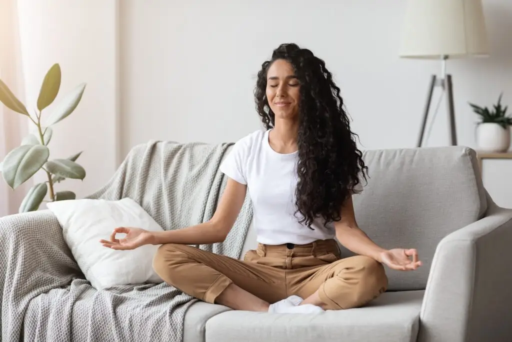 Young brunette woman with long curly hair sitting cross-legged on her couch in a meditation pose. Her arms are resting on her legs and her thumbs are touching her middle fingers.