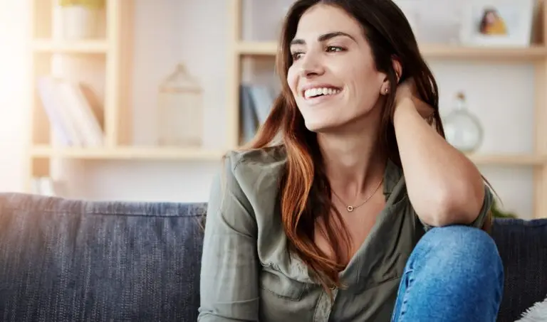 Brunette woman sitting on her couch with one knee up, elbow resting on her knee, looking off to the side smiling because she learned how to practice mindfulness.