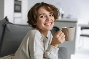 Young, short-haired brunette woman holding her coffee mug while perched up on her couch, smiling because she has used progressive muscle relaxation and reduced her anxiety and depression symptoms.