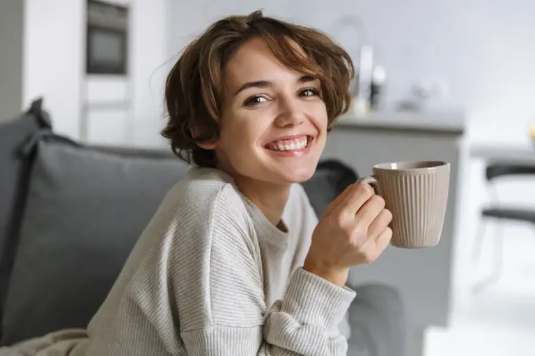 Young, short-haired brunette woman holding her coffee mug while perched up on her couch, smiling because she has used progressive muscle relaxation and reduced her anxiety and depression symptoms.