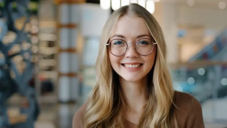 A close up of a young blonde woman wearing glasses and smiling wide because she learned how to reduce her anxiety symptoms with progressive muscle relaxation.