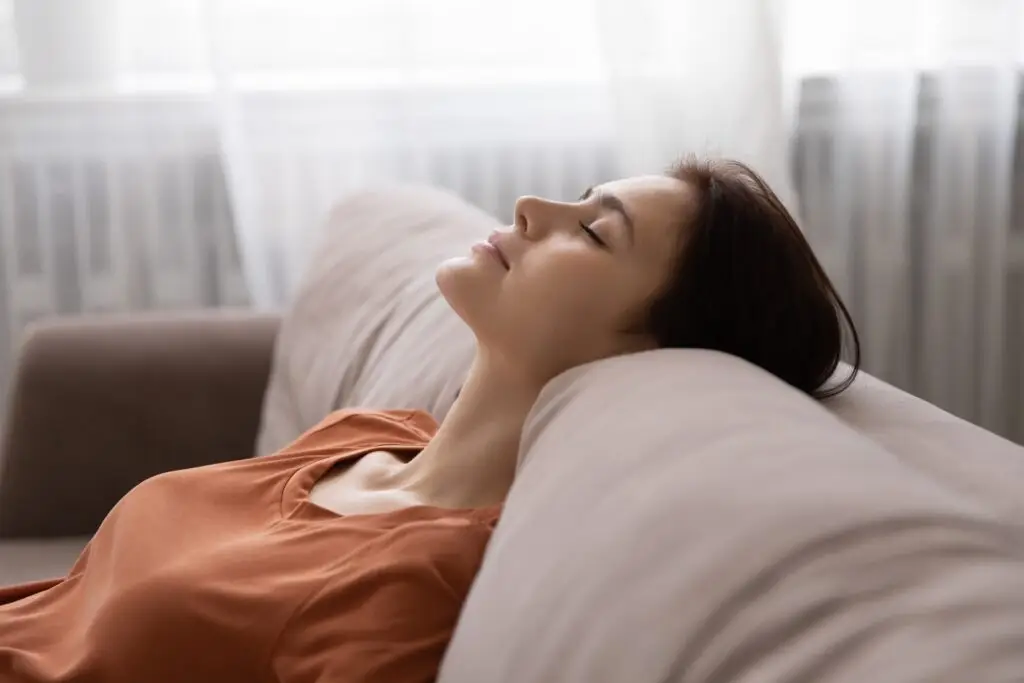 A young brunette woman is sitting on her couch with her head leaning back onto the cushion of the couch, eyes closed, using the different progressive muscle relaxation techniques she has learned to reduce stress.