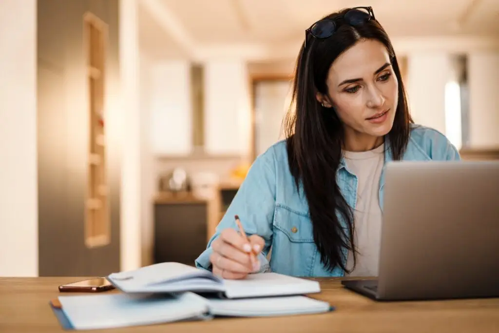 A woman writing down the steps to practice radical acceptance in a notebook while she does online research.