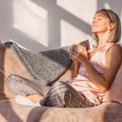 Happy dreamy middle aged woman sitting on sofa in living room with cup of tea or coffee