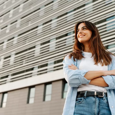 woman standing outside of her workplace in jeans and a long sleeved buttoned shirt, feeling confident and looking off into the distance