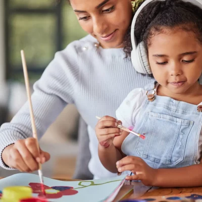 A mother has her young daughter sitting in her lap as they both paint. The young girl has headphones on to reduce sensory input, as she has autism and is sensitive to sounds.