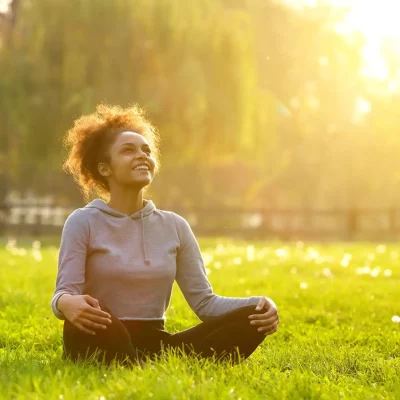 young black woman sitting in the grass at a part in lotus pose, legs crossed, sun shining through the trees as she smiles