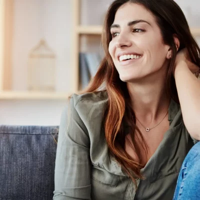 Brunette woman sitting on her couch with one knee up, elbow resting on her knee, looking off to the side smiling because she learned how to practice mindfulness.