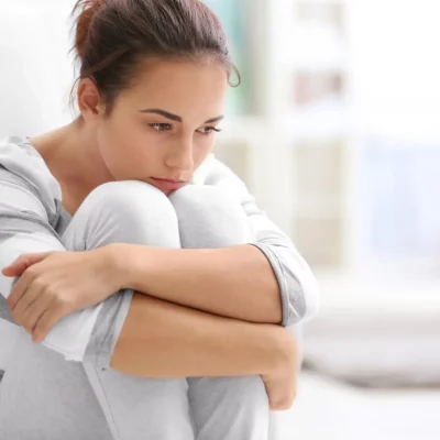 A young woman holding her legs into her chest, arms around her knees distressed about her binge eating disorder.