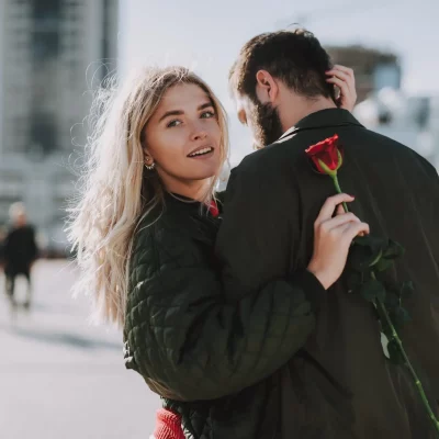 A young woman embracing her significant other while holding a rose behind his back.