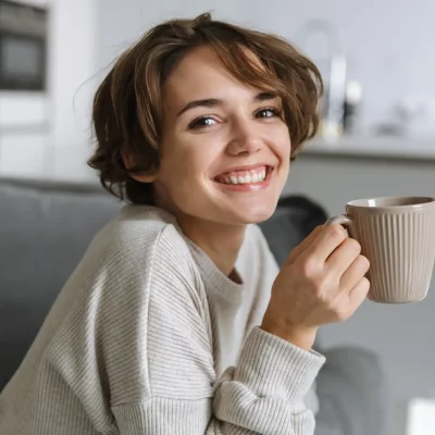 Young, short-haired brunette woman holding her coffee mug while perched up on her couch, smiling because she has used progressive muscle relaxation and reduced her anxiety and depression symptoms.