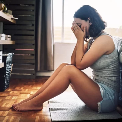 young brunette woman sitting on her floor, elbows resting on her knees, hands covering her face due to stress and the job market