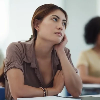 Young woman in a class feeling distracted, looking off to the side while her classmates are focused on their computers doing work.